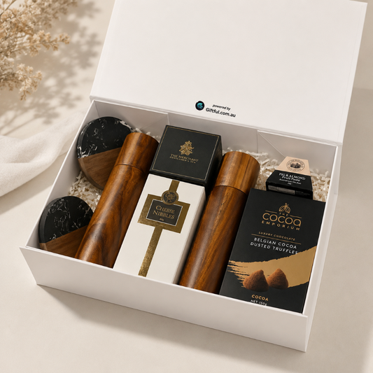 An open white gift box containing a curated chocolate and gift set, including a boxed cheese wedge, two wooden-handled items, and a box of cocoa dusted truffles, arranged on a light neutral surface with a soft decorative cloth and dried flowers nearby.