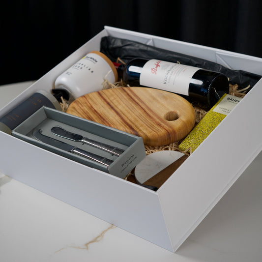 Gift box with wine bottles, cutting board, and utensils on a marble surface.