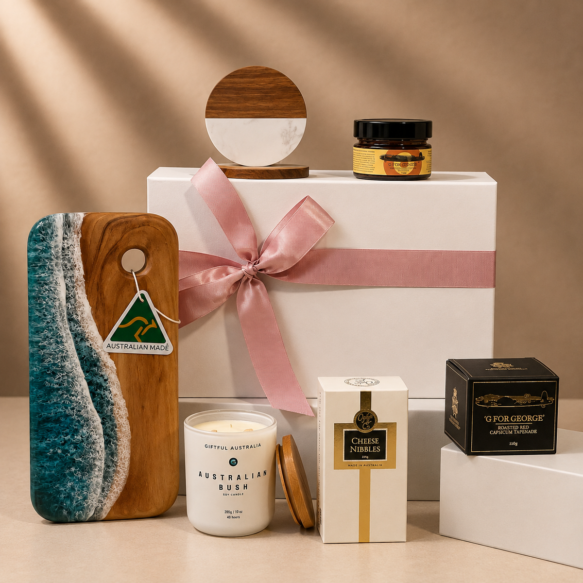 A curated gift set arranged on a beige surface against a warm beige backdrop. The set includes a white gift box tied with a pink ribbon, a wooden board with a turquoise and white wave design, a white candle labeled 'Australian Bush,' a cream box labeled 'Chilem Ambre,' a black box labeled 'Gourge Geologique,' a small jar with a gold label, and a round wooden-and-white decor piece.