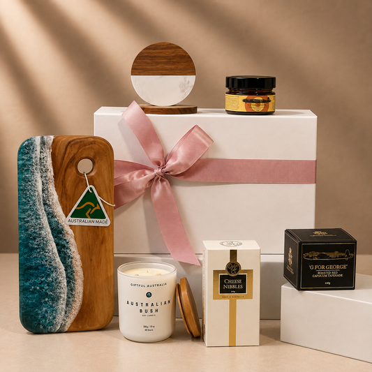 A curated gift set arranged on a beige surface against a warm beige backdrop. The set includes a white gift box tied with a pink ribbon, a wooden board with a turquoise and white wave design, a white candle labeled 'Australian Bush,' a cream box labeled 'Chilem Ambre,' a black box labeled 'Gourge Geologique,' a small jar with a gold label, and a round wooden-and-white decor piece.