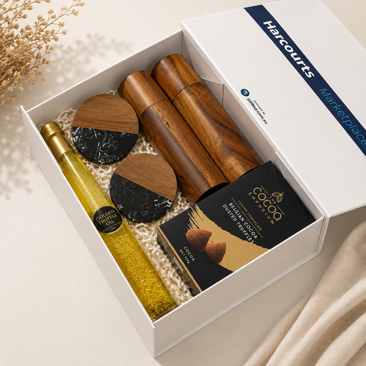 A luxury gift box from Harcourts Marketplace containing a bottle of olive oil, two wooden and marble salt/pepper mills, a chocolate-covered nut spread, and a box of Harcourts Belgian Chocolate Duttweiler, arranged in white crinkle paper on a beige background.