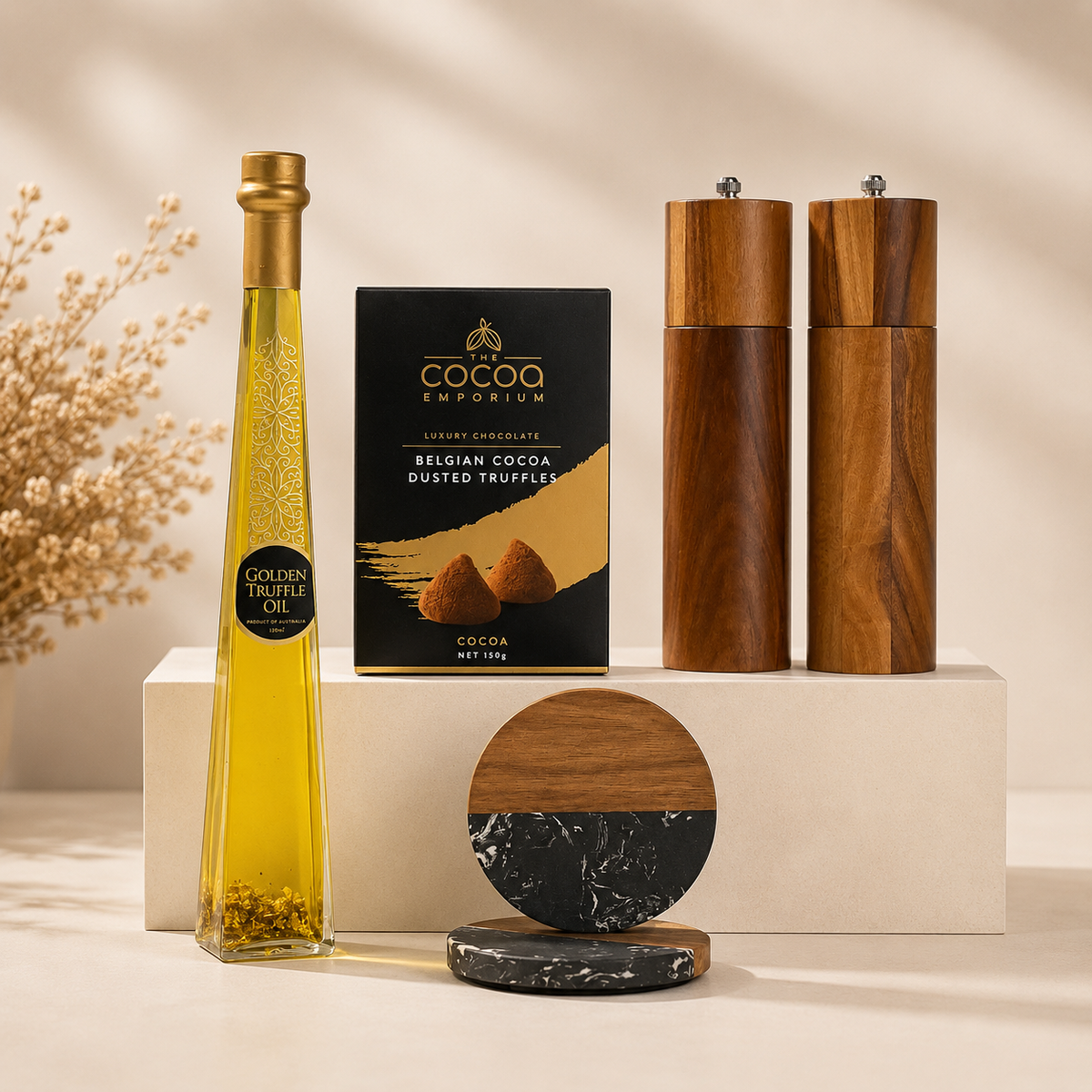 A styled product arrangement on a cream pedestal featuring a bottle of golden truffle oil, a box of Cocoa Emperium Belgian Cocoa Dusted Truffles, two wooden pepper and salt mills, and two round coasters with a wood-and-marble design.