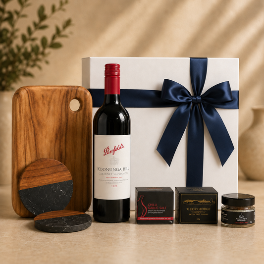A luxury gift hamper set featuring a bottle of Penfolds Koonunga Hill red wine, a white gift box with a navy blue ribbon, a wooden cutting board, two round coasters, and gourmet food boxes on a beige surface.