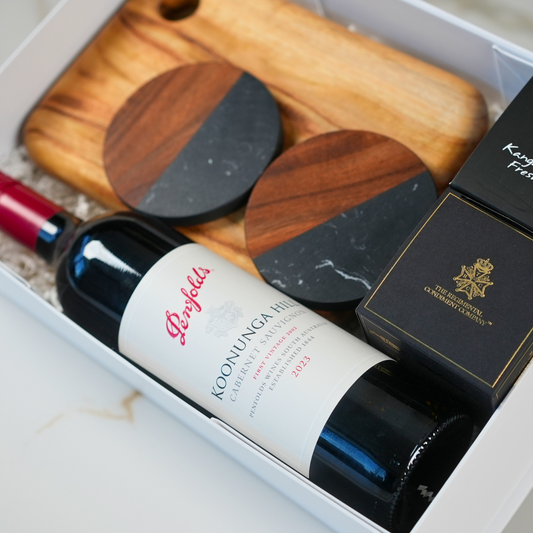 A gift set containing a bottle of Penfolds Koonunga Hill Cabernet Sauvignon 2023, two marble and timber coasters, a wooden serving board, and a box of luxury artisan chocolates.