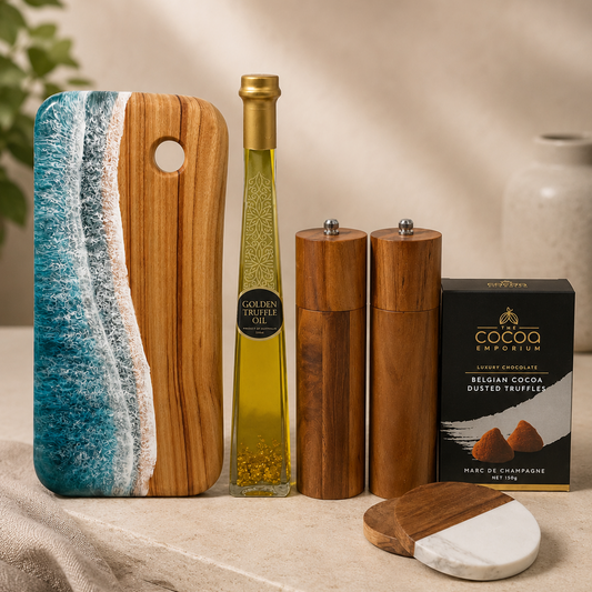 A styled gift set on a stone surface featuring a wooden cutting board with a blue ocean wave design, a bottle of olive oil, two wooden salt and pepper mills, a box of Cocoa Lusso truffles, and a marble-and-wood cheese knife.