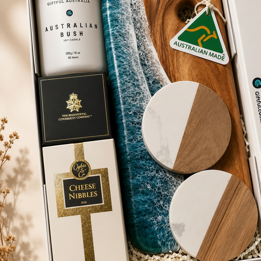 An open gift box arranged with Australian-made products: a white candle labeled 'Australian Bush' candle, a black and gold box, a white and gold 'Cheese Nibbles' box, a blue-and-teal textured cloth, a wooden board, two round coasters with white and wood halves, and a red-and-white box. The scene is styled on a beige surface with dried flowers at the side.